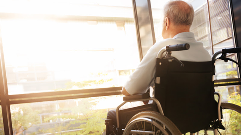 Senior Man in Wheelchair looking out of a window in a hospital corridor.